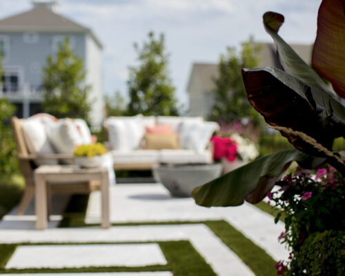 Modern backyard with a wooden bench, outdoor cushions, and potted plants; the foreground features large green leaves and flowers—perfect for relaxing or entertaining in an outdoor living space.