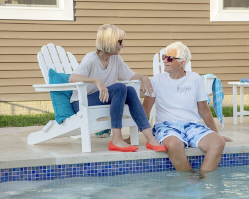 Two older adults sit on white chairs by an inground pool, feet in the water, chatting and wearing sunglasses. A towel and pool toys are visible in the background.