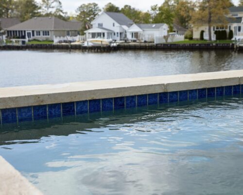 A close-up view of a luxury pool edge with water, overlooking a calm waterfront neighborhood with houses and docks in the background—perfect for refined outdoor living.