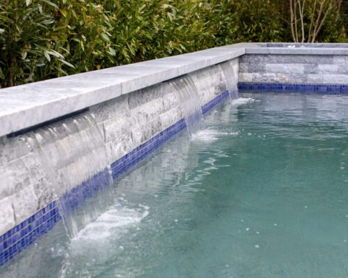 Rectangular inground pool with water cascading from three stone spouts along the edge, surrounded by lush green shrubs—perfect for refined outdoor living.