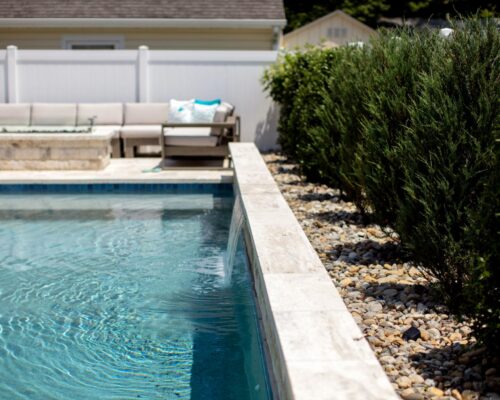A backyard luxury pool with clear water, a stone border, a row of shrubs on the right, and outdoor seating with cushions in the background.
