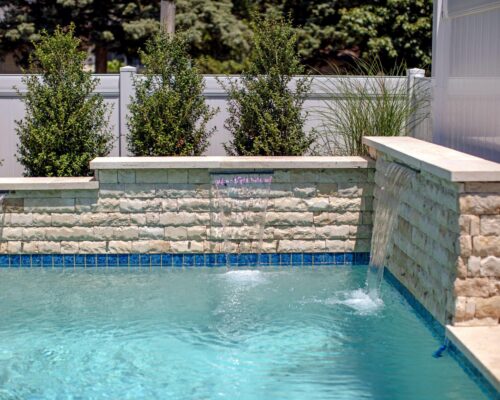 A luxury pool with water features flowing from a stone wall, bordered by shrubs and a white privacy fence, creates the perfect outdoor living retreat.