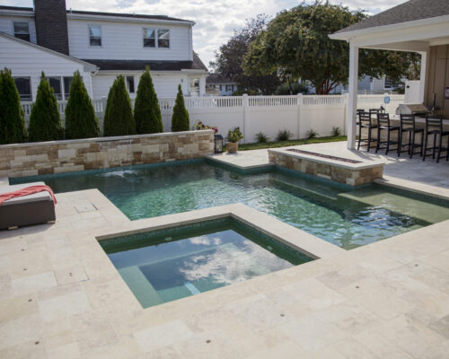 Rectangular inground pool with attached hot tub, surrounded by light stone tiles, outdoor seating, and a covered bar area. White fence and shrubs line the backyard, creating the perfect setting for custom spas or fiberglass pools.