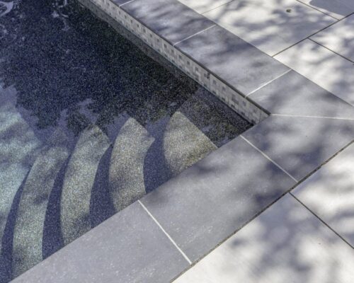 Shallow steps lead into a tiled luxury pool, with sunlight and shadows cast on the surrounding light-colored pool deck—perfect for pairing with custom spas or outdoor kitchens.
