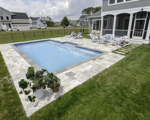 A rectangular luxury pool with a surrounding stone patio, outdoor seating, and potted plants sits adjacent to a gray house with a screened porch—perfect for refined outdoor living.