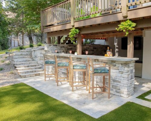 Outdoor living at its best with a stone bar, four wooden barstools, and drinks on the counter, all beneath a wooden deck in a landscaped backyard—perfect for relaxing after a swim in nearby fiberglass pools.