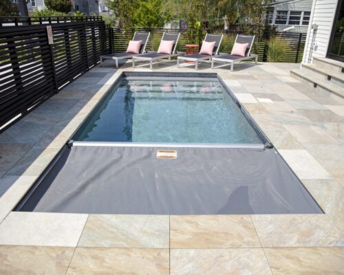 A small rectangular fiberglass pool with a partially covered safety cover is surrounded by stone tile, with lounge chairs and pillows in the background—perfect for relaxing outdoor living.