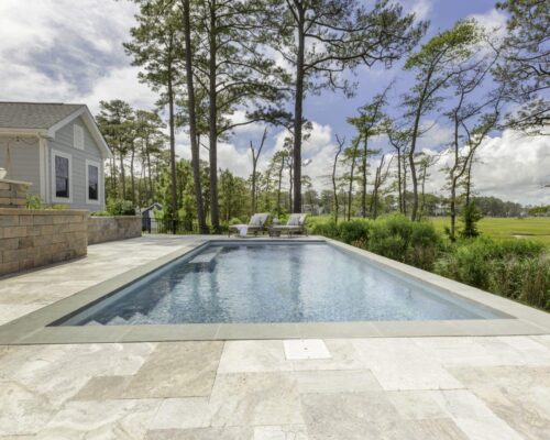 Rectangular luxury pool with stone tile deck, two lounge chairs at the far end, surrounded by tall trees and greenery, next to a house under a partly cloudy sky—perfect for outdoor living.