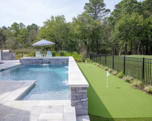A backyard featuring an inground pool on the left and a small artificial putting green with two flags on the right, separated by a black metal fence.