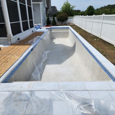 An empty inground pool under construction beside a house, surrounded by white fencing and partially covered with plastic sheets—an inviting addition to enhance your outdoor living space.