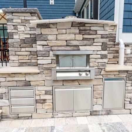 Built-in outdoor kitchen with a stone facade, featuring a stainless steel grill, oven, storage drawers, and cabinets set against a tiled patio—perfect for entertaining near custom spas or luxury pools.