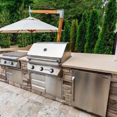 Outdoor kitchen with stone countertops featuring a gas grill, side burner, mini fridge, and umbrella, set on a stone patio with trees and a house in the background—perfect for homes with outdoor kitchens or luxury pools.