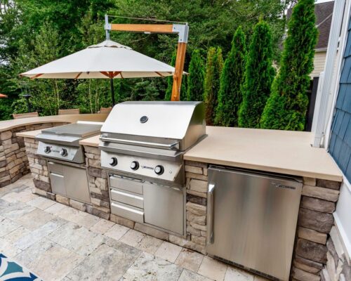 Outdoor kitchen with stone countertops featuring a gas grill, side burner, mini fridge, and umbrella, set on a stone patio with trees and a house in the background—perfect for homes with outdoor kitchens or luxury pools.