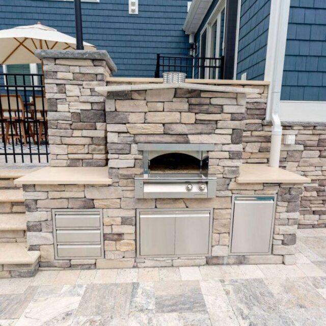 Outdoor built-in stone grill and oven with stainless steel cabinets, set against a stone patio and blue house exterior—an ideal centerpiece for luxury pools and stunning outdoor kitchens.