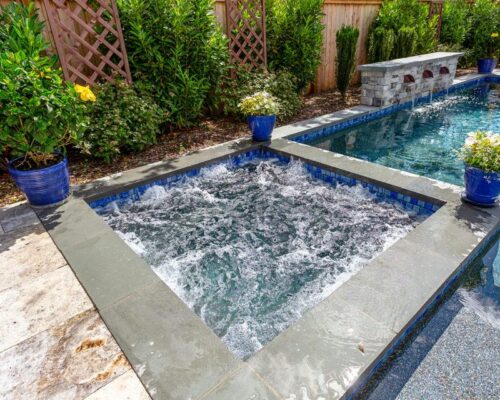 A bubbling hot tub adjacent to a luxury pool, surrounded by potted plants and greenery in a fenced backyard, creates the perfect outdoor living retreat.