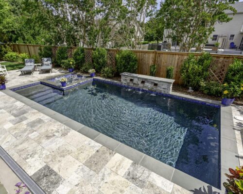 Rectangular backyard fiberglass pool with clear water, stone tile deck, lounge chairs, potted plants, and a wooden privacy fence with landscaping—perfect for elevated outdoor living.