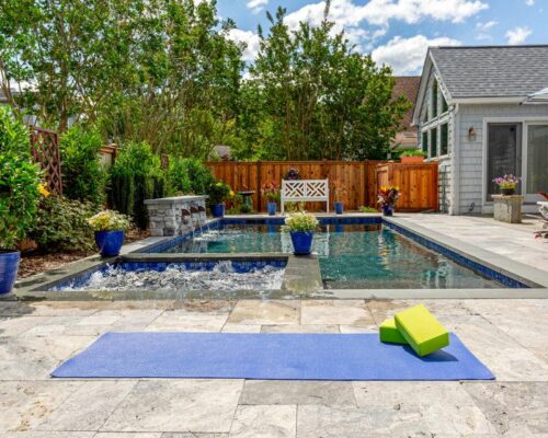 A backyard designed for outdoor living, featuring a rectangular inground pool, potted plants, a small waterfall feature, and a patio area with a blue yoga mat and two green yoga blocks on the stone paving.