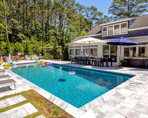 A backyard with a rectangular inground pool, lounge chairs, umbrellas, and a modern house surrounded by trees on a sunny day—perfect for relaxing or enjoying meals from nearby outdoor kitchens.