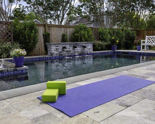 A backyard inground pool with a stone fountain is surrounded by greenery. A purple yoga mat and two green blocks rest on the stone patio, creating an inviting outdoor living space beside the pool.