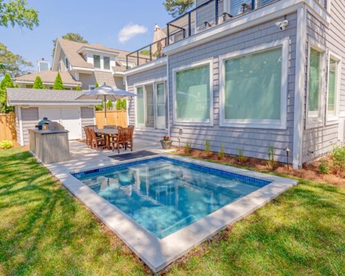 A small backyard features a rectangular luxury pool, patio with dining table and chairs, an outdoor grill, and a grey-sided house with large windows.