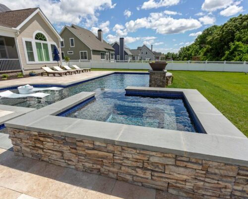 Modern backyard with a stone-edged hot tub connected to a rectangular fiberglass pool, surrounded by patio chairs and grass, creating an inviting outdoor living space with homes and a white fence in the background.