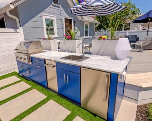 Outdoor kitchen with blue cabinets, built-in grill, sink, and mini fridge on a patio with lounge seating and umbrellas—perfect for outdoor living—adjacent to a gray house.