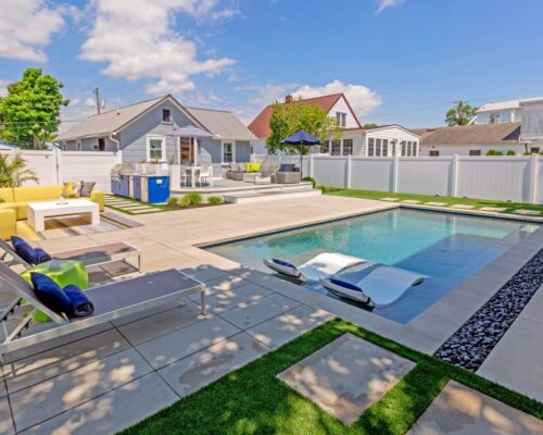 Modern backyard with a rectangular swimming pool, lounge chairs, and an outdoor seating area surrounded by white fencing and landscaped greenery—ideal for outdoor living and relaxing in style.