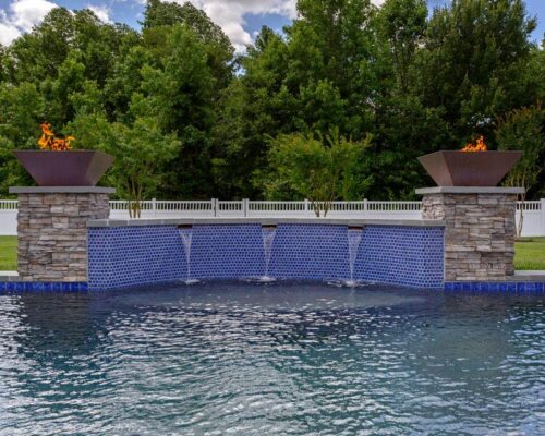 A luxury inground pool features three water spouts flowing from a stone wall, topped with two fire bowls. Trees and a white fence in the background complete this stunning outdoor living space.