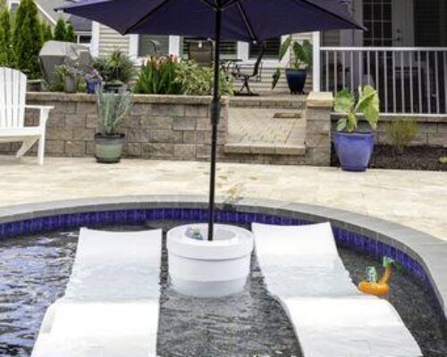 Two white lounge chairs and a small table with a blue umbrella are set up on a shallow ledge in a luxury pool; a toy duck floats nearby.