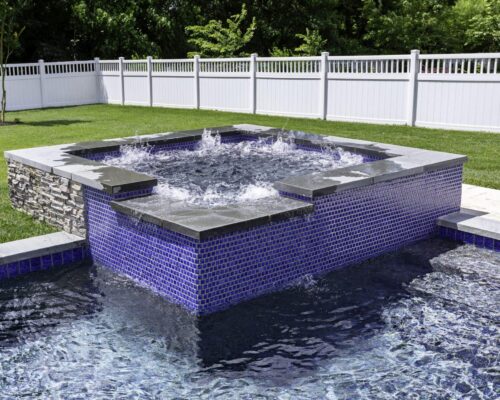 Rectangular custom spa with blue tiles and stone trim overflows into a luxury pool in a fenced backyard with green grass, trees, and space for outdoor kitchens.