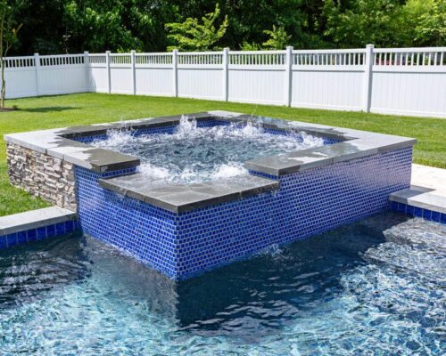 A backyard pool designed for outdoor living, with a raised square hot tub featuring blue tile and stone accents, surrounded by grass and a white fence.