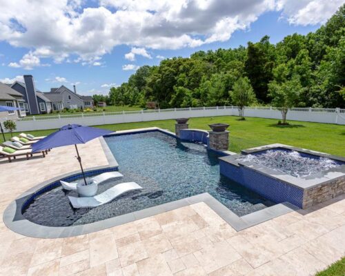 Backyard inground pool with attached hot tub, lounge chairs, and umbrella set on a stone patio, surrounded by grass, trees, and a white fence under a partly cloudy sky—perfect for enjoying luxury pools at home.