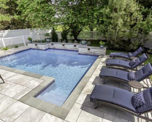 Rectangular backyard luxury pool with four lounge chairs on a tiled patio, surrounded by greenery and a white fence—the perfect setting to complement outdoor kitchens or custom spas.