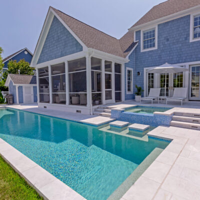 Backyard view of a modern house with blue siding, screened porch, outdoor seating, a long inground pool, and a raised hot tub under a clear sky. An inviting space perfect for relaxing or entertaining near stylish outdoor kitchens.