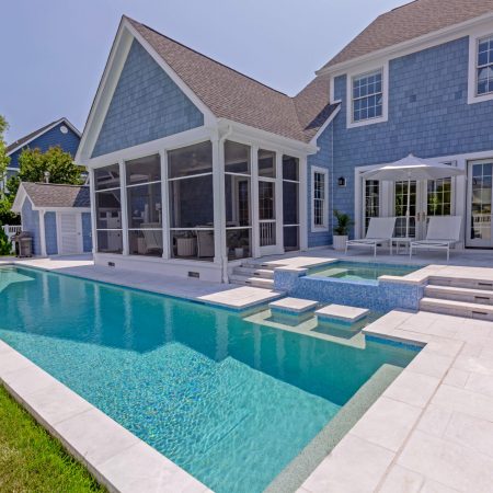 Backyard view of a modern house with blue siding, screened porch, outdoor seating, a long inground pool, and a raised hot tub under a clear sky. An inviting space perfect for relaxing or entertaining near stylish outdoor kitchens.