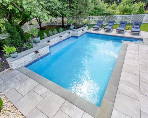 Rectangular inground pool with light stone tile deck, adjacent lounge chairs, potted plants, and surrounding greenery, all enclosed by a white fence—perfect for relaxed outdoor living.
