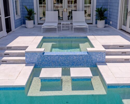 A small raised custom spa with blue tile overflows into a rectangular inground pool, surrounded by white lounge chairs, potted plants, and light-colored tile steps.