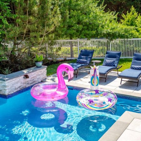 A backyard oasis featuring a fiberglass pool with two colorful inflatable floats, surrounded by lounge chairs and potted plants—perfect for outdoor living, all enclosed by a white fence and green trees.
