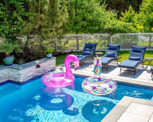 A backyard oasis featuring a fiberglass pool with two colorful inflatable floats, surrounded by lounge chairs and potted plants—perfect for outdoor living, all enclosed by a white fence and green trees.
