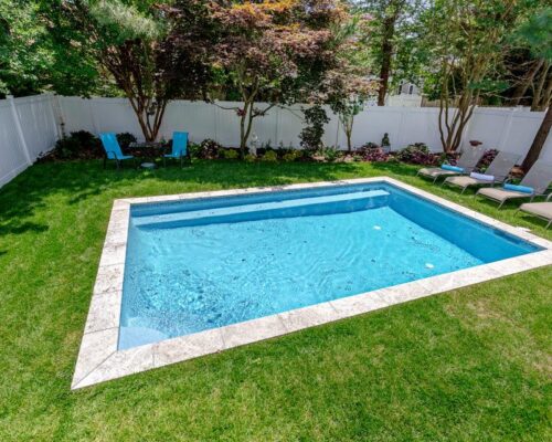 Rectangular inground pool surrounded by grass, lounge chairs, and a white fence, with trees and plants along the fence line—perfect for outdoor living.