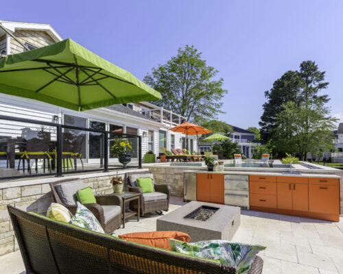Modern outdoor patio with wicker seating, a fire pit, stainless steel outdoor kitchen area with orange cabinets, and large green and orange umbrellas under a clear blue sky—perfect for relaxing beside inground pools.