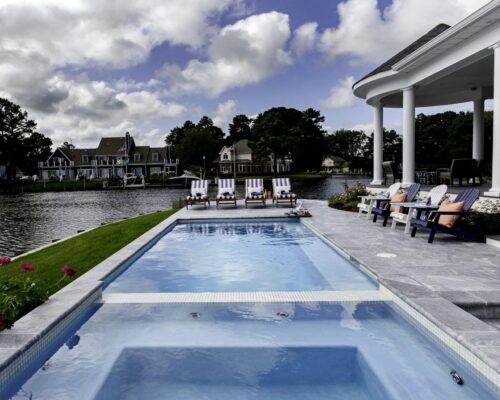 A rectangular inground pool with adjacent jacuzzi, lined with lounge chairs, sits beside a waterfront home under a partly cloudy sky.