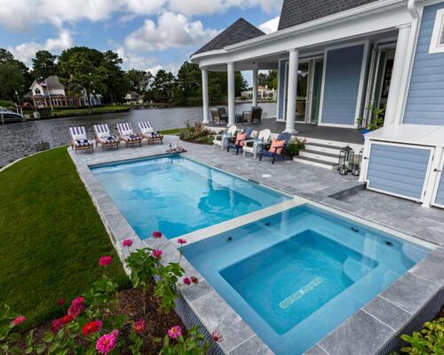 A modern house with a fiberglass pool and hot tub in the backyard, surrounded by lounge chairs, offers exceptional outdoor living and overlooks a lake with trees and houses in the background.