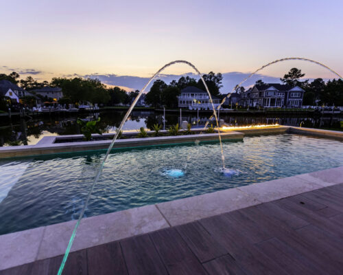 Modern backyard luxury pool with two arched water jets at sunset, surrounded by landscaped plants, a canal, and houses in the background—perfect for enjoying beside outdoor kitchens.