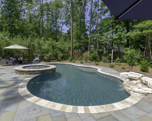 Backyard with a curved inground pool, attached custom spa, lounge chairs under umbrellas, stone patio, and trees in the background.
