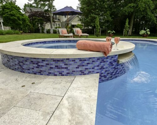 A round hot tub with blue mosaic tiles overflows into a swimming pool, creating the perfect outdoor living retreat. A rolled towel and two glasses rest on the edge; lounge chairs and trees complete this inviting scene.