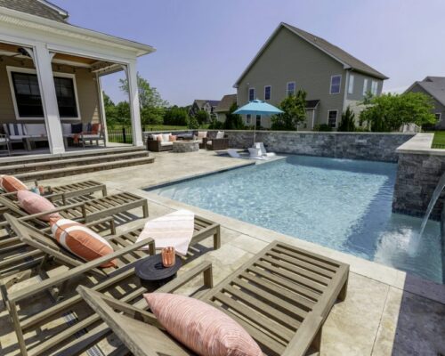 Outdoor patio with wooden lounge chairs and pillows beside a rectangular inground pool, adjacent to a covered seating area, with houses and greenery in the background.