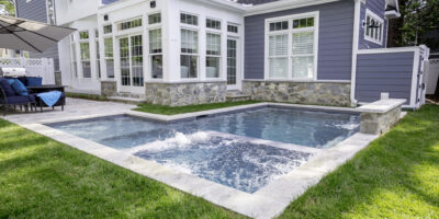 A modern backyard featuring a small inground pool and attached hot tub, surrounded by grass and a gray house with large windows—perfect for relaxing or enhancing your outdoor living experience.