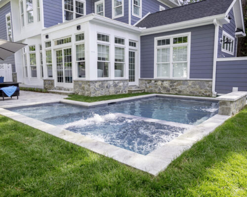 A modern backyard featuring a small inground pool and attached hot tub, surrounded by grass and a gray house with large windows—perfect for relaxing or enhancing your outdoor living experience.