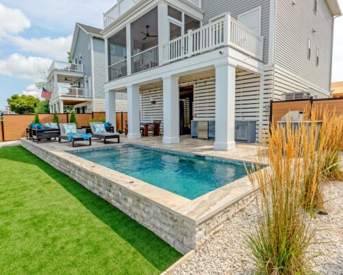 Modern backyard with a rectangular luxury pool, patio seating, artificial grass, and a two-story house featuring covered balconies and wood fencing—perfect for stylish outdoor living.